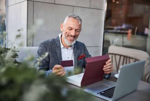Joyous aging man looking at a dark-red card in his hand and smiling while sitting at a cafe